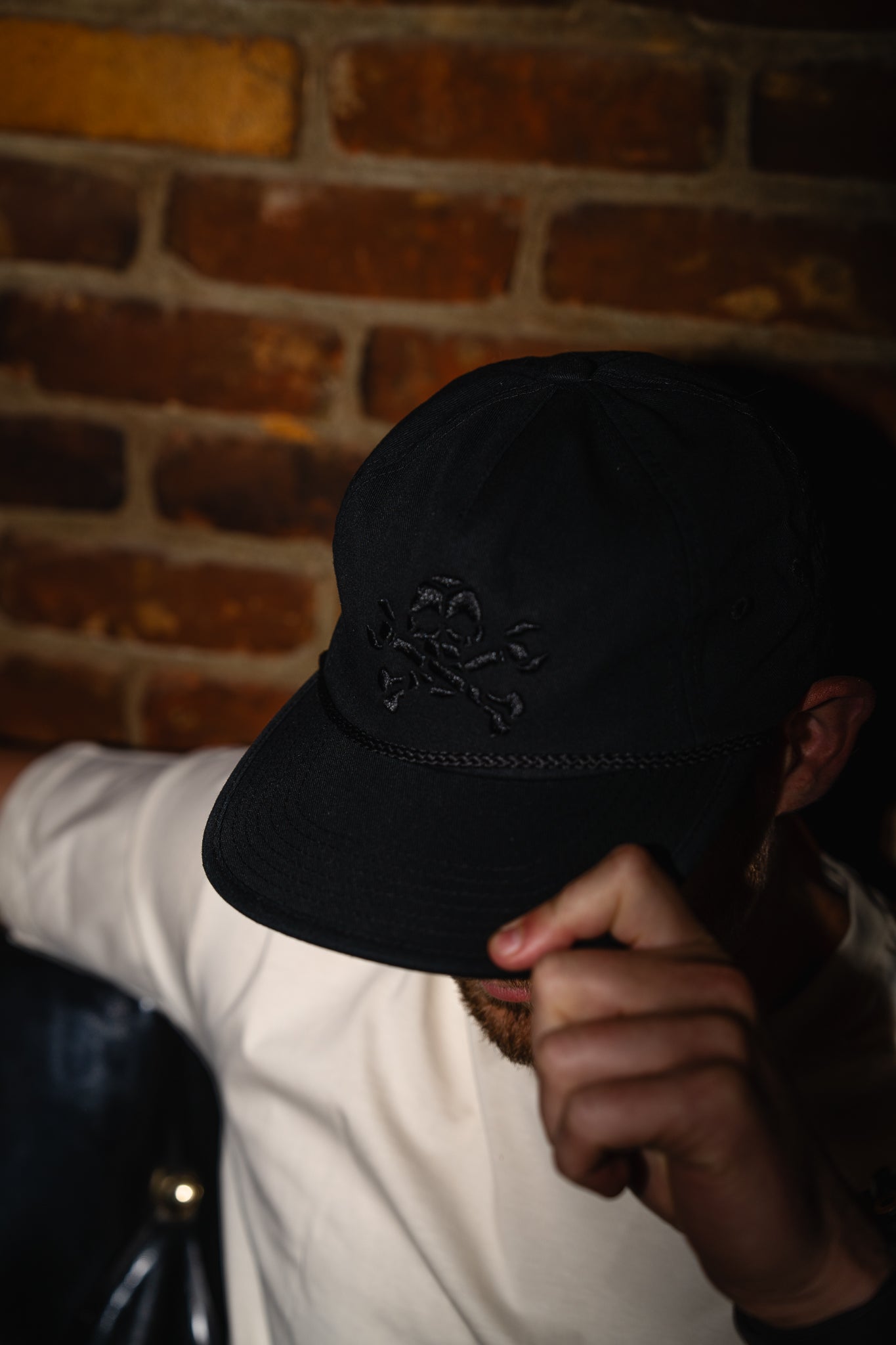 Person wearing a black cap with a logo against a brick wall.