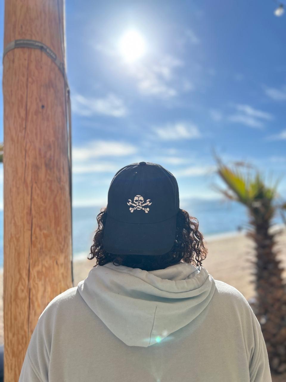 man standing on a beach wearing a backwards black dad hat with a Boneyard skull and cross bones