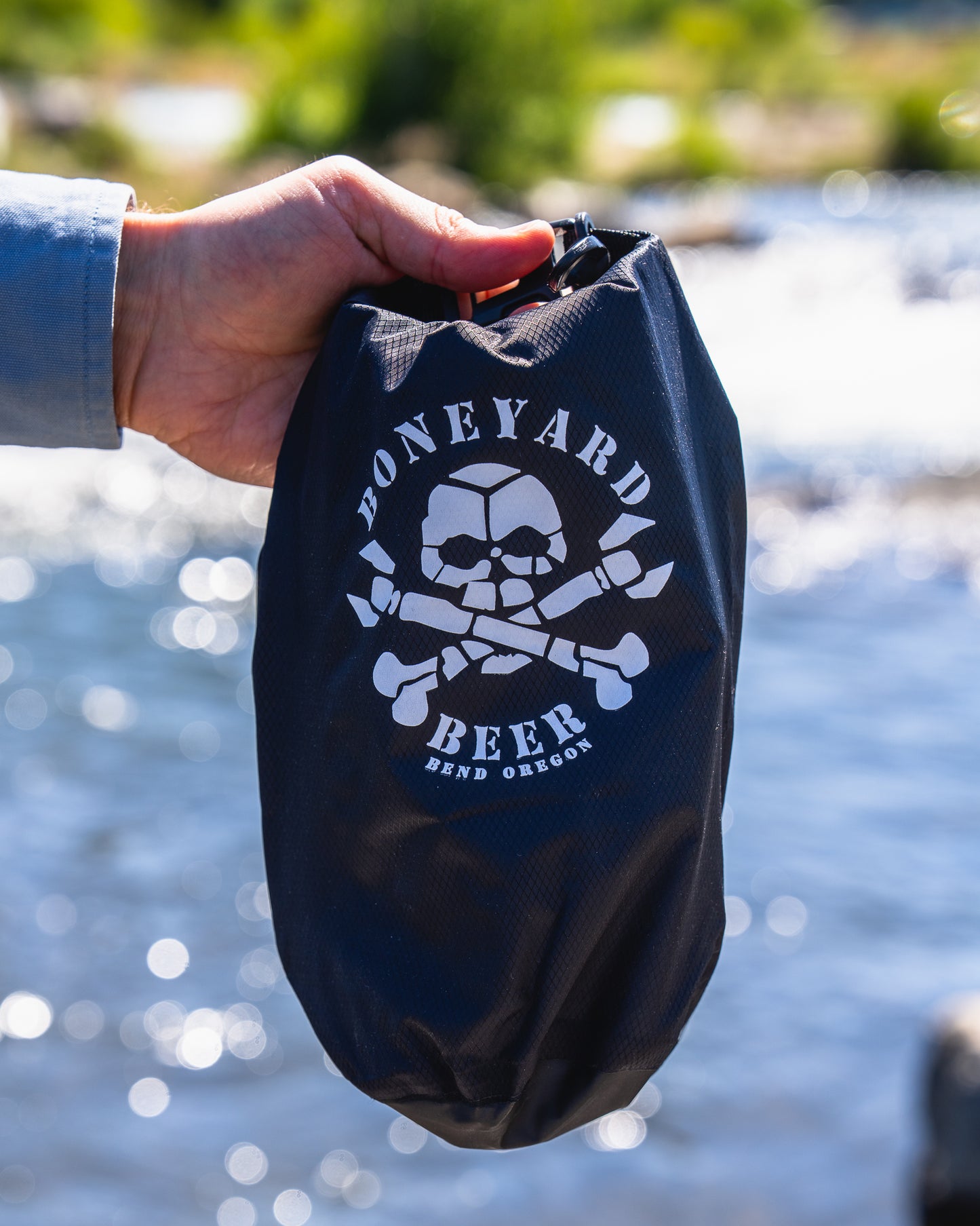 Hand holding a black bag with 'Boneyard Beer' logo against a blurred natural background