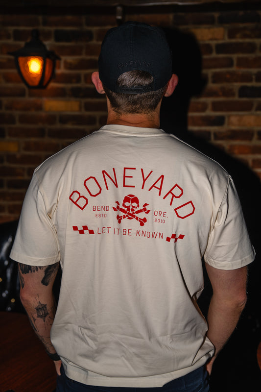 Person wearing a beige shirt with 'Boneyard' logo in red on the back, standing against a brick wall.