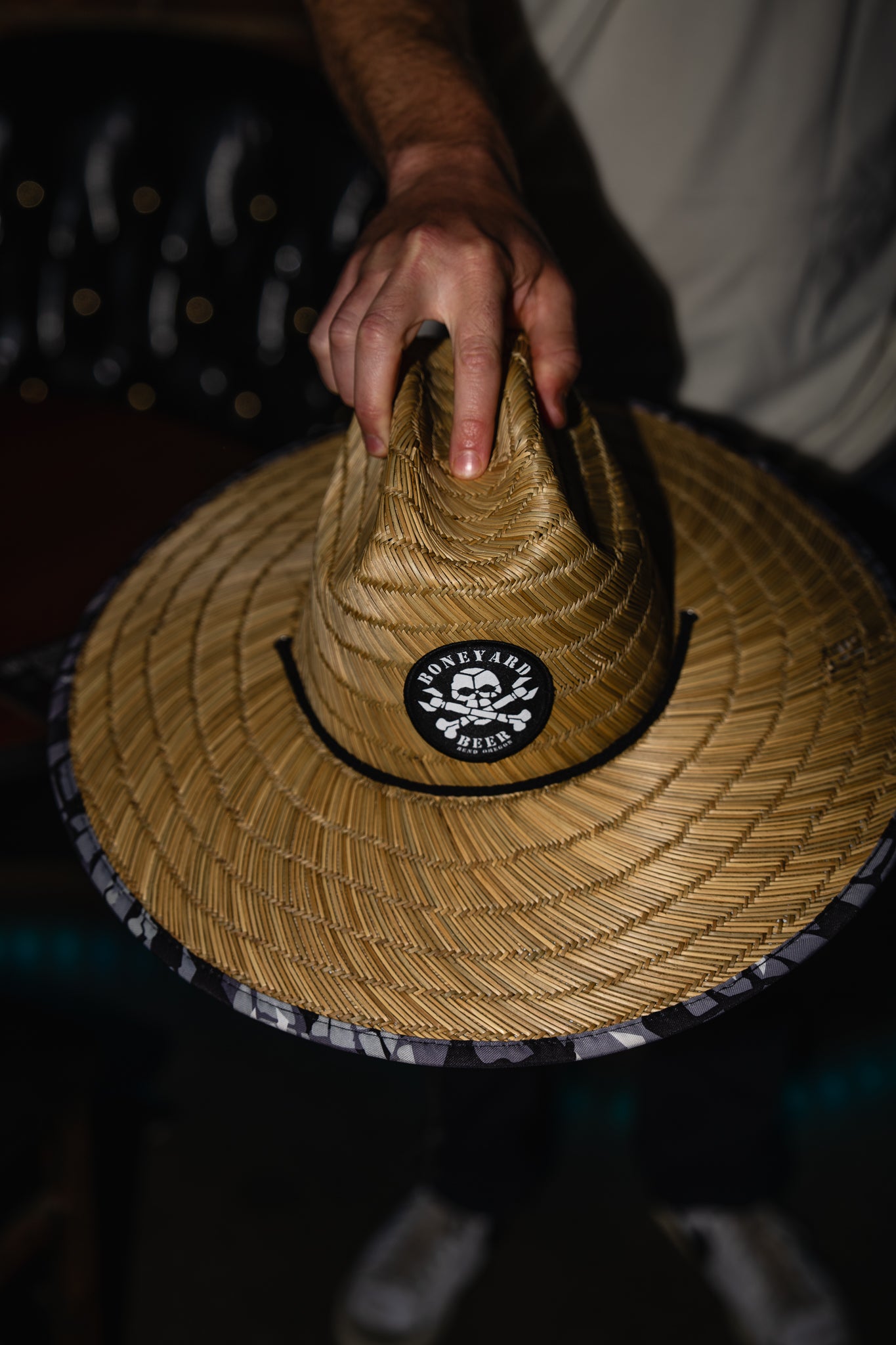 Hand holding a straw hat with a logo on a dark background