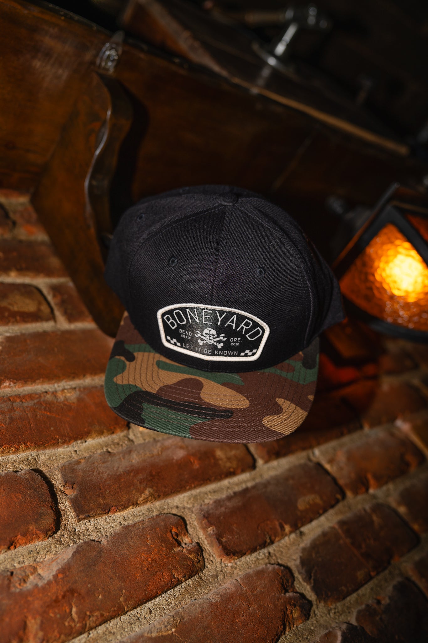 Black cap with 'Boneyard' logo and a camo bill on a brick surface