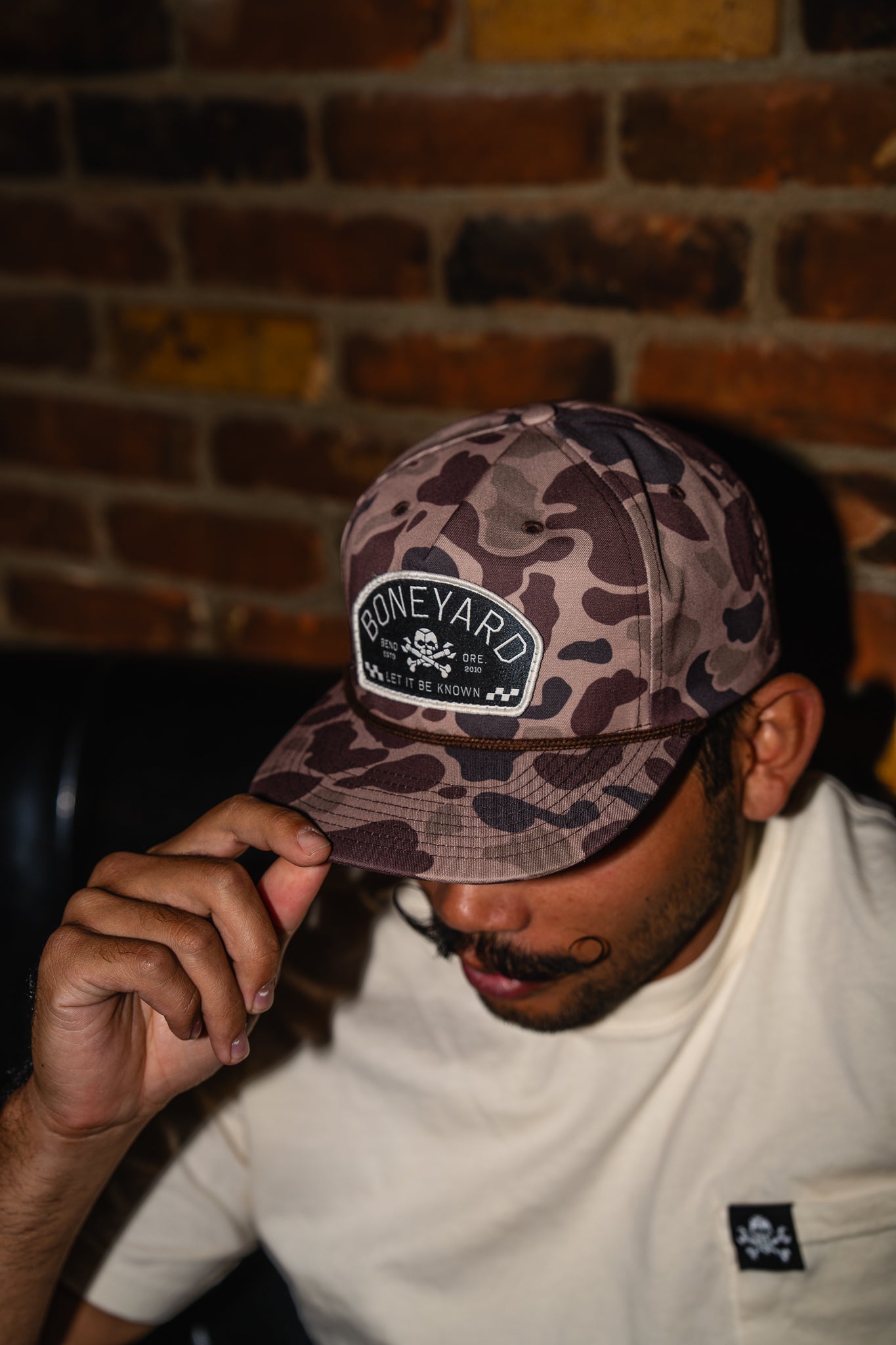 Person wearing a camouflage cap with 'Boneyard' logo against a brick wall.
