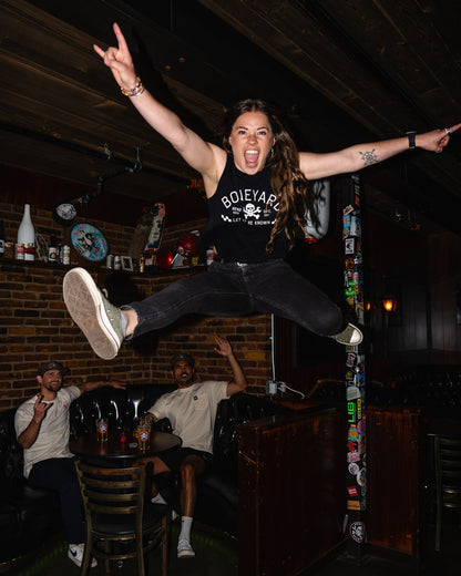 Person jumping in a bar with friends, wearing a black t-shirt with 'Boileyard' text.