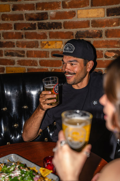 Man sitting in a booth with a brick wall background, holding a beer glass wearing a boneyard beer hat