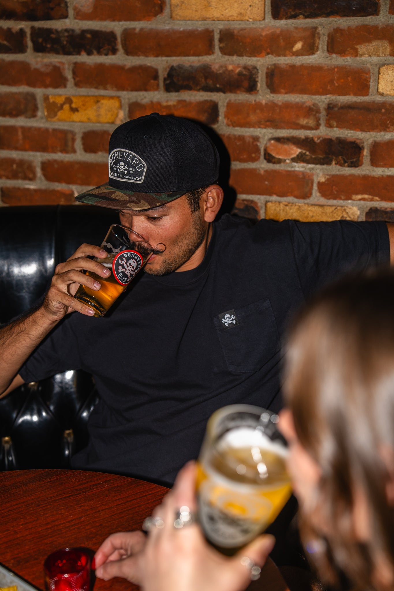 Man drinking a beer in a bar with a brick wall background wearing a block boneyard beer hat with a camo bill.
