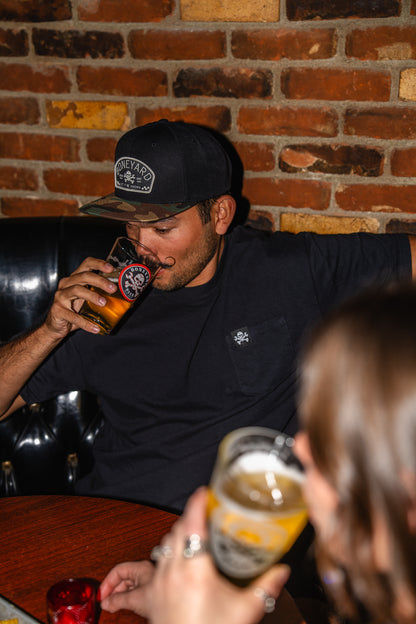 Man drinking a beer in a bar with a brick wall background wearing a block boneyard beer hat with a camo bill.