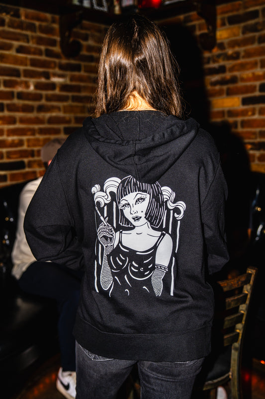 Person wearing a black hoodie with a graphic design on the back, standing in a dimly lit room with brick walls.